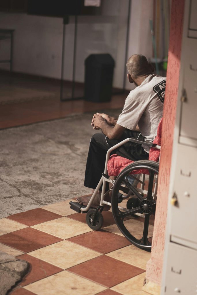 A man in a wheelchair sits alone indoors, reflecting quietly in a warm-toned environment.